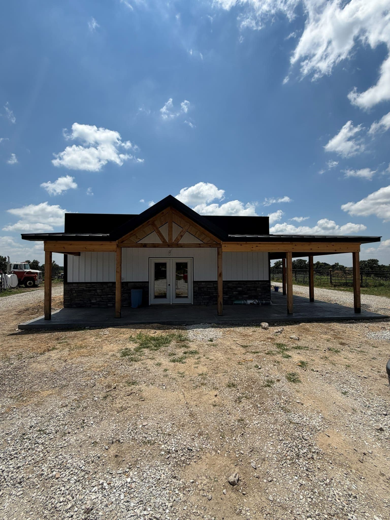 Front gable of a post-frame building with a cedar timber-truss porch, stone wainscot and black standing-seam roof